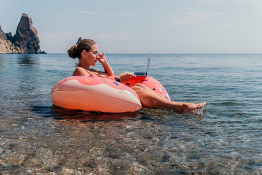 Woman freelancer works on laptop swimming in sea on pink inflatable ring. Pretty lady typing on computer while floating in the sea on inflatable donut at sunset. Freelance, remote work on vacation