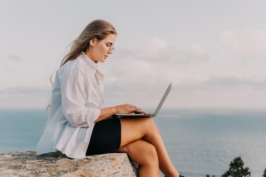 Digital nomad, woman in the hat, a business woman with a laptop sits on the rocks by the sea during sunset, makes a business transaction online from a distance. Freelance, remote work on vacation.