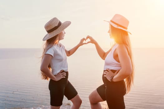 Two women wearing straw hats stand on a cliff overlooking a body of water as they make a heart shape with their hands. The sunset illuminates the scene with a warm glow.