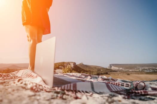A woman in a red dress stands on a mountaintop with a laptop on a checkered blanket in front of her.