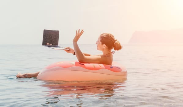 Woman freelancer works on laptop swimming in sea on pink inflatable ring. Pretty lady typing on computer while floating in the sea on inflatable donut at sunset. Freelance, remote work on vacation
