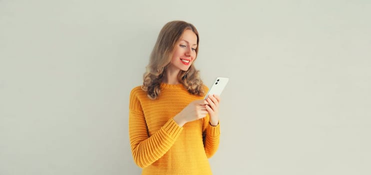 Portrait of happy smiling young woman looking at smartphone in yellow sweater on gray background