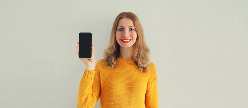 Portrait of happy smiling young woman showing black blank smartphone screen on gray background