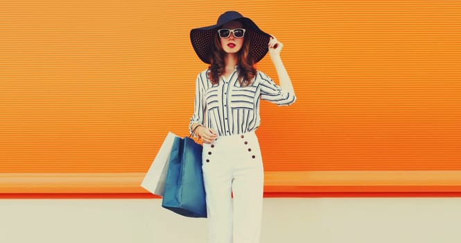Beautiful young woman with shopping bags in white striped shirt, summer hat on orange background