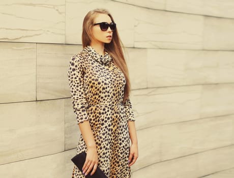 Portrait of beautiful blonde young woman model wearing leopard dress, handbag clutch and sunglasses on wall background