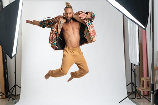 Full length gay man jumping wearing stylish clothing dancing on white studio background. Brazilian homosexual male posing in photo studio. Bearded Afro-American gay man cut out on white.