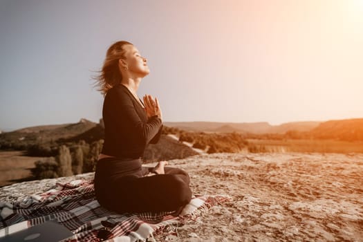 A woman in black clothing sits in a meditative pose on a rocky mountaintop, overlooking a valley. She has a plaid blanket and a laptop beneath her, suggesting a blend of nature and technology.
