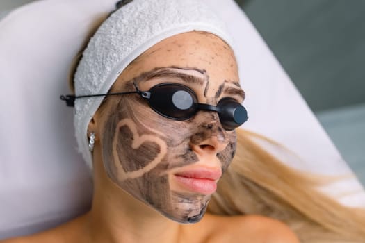 cosmetologist applying black mask on pretty woman face wearing black gloves, gorgeous woman in spa having facial procedures.