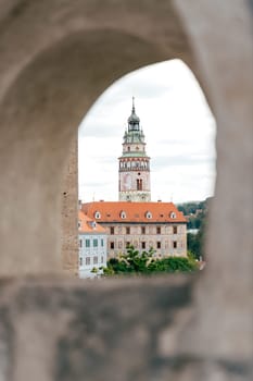 The view showcases a historic tower adorned with a clock, standing prominently against a backdrop of a quaint European town. The tower features a distinctive roof and is framed by a weathered stone archway.
