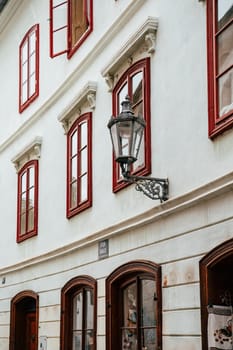 This location features a beautiful historic building adorned with striking red windows and a vintage lantern. The architecture reflects classic European design elements, inviting viewers to explore the quaint street where the building is situated.
