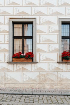 Bright red geraniums bloom in wooden flower boxes beneath two windows, framed by a textured wall adorned with geometric patterns. The setting is a quaint European town, showcasing beautiful architectural details.
