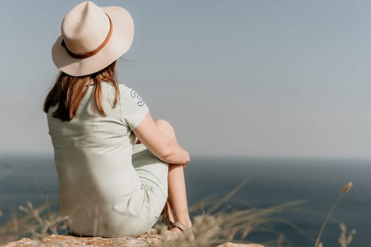 A woman wearing a straw hat sits on a cliff, looking out at the vast, blue ocean in a contemplative pose.