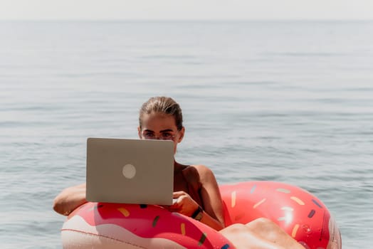 Woman freelancer works on laptop swimming in sea on pink inflatable ring. Pretty lady typing on computer while floating in the sea on inflatable donut at sunset. Freelance, remote work on vacation