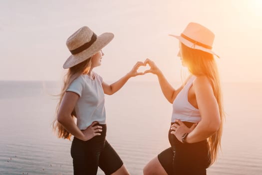 A mother and daughter stand facing each other on a cliff overlooking a body of water. They are wearing casual clothing and both have their hands up forming a heart shape.