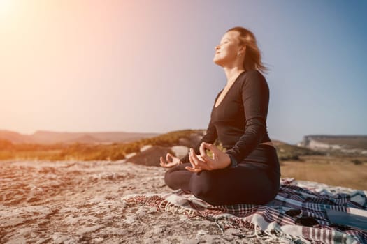 A woman is meditating in a peaceful pose while sitting on a rock with a blanket on a mountain top with a vast nature view.