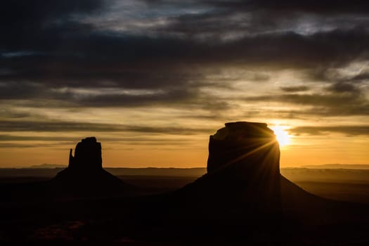 Breathtaking sunrise over Monument Valley, Arizona, featuring dramatic silhouettes of the iconic Mittens rock formations with lens flare. This landscape captures the beauty of the American Southwest.
