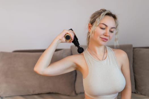 Girl at home holding a therapeutic percussion vibrating massage gun in her hand and massaging her back muscles Sports recovery concept after a workout. . High quality photo