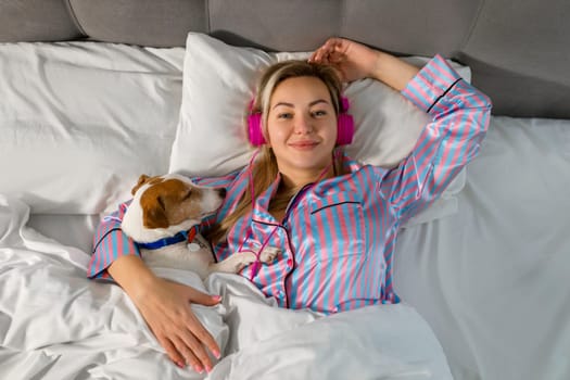 Carefree lady in pajamas listening to music on headphones while sitting in bed with her beloved pet.