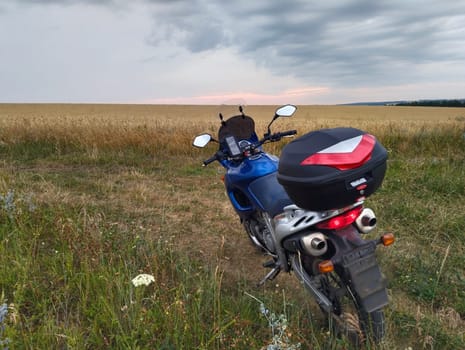 A motorcycle rests on the grass in an open field, surrounded by tall grass and under a dramatic sky as the sun sets.