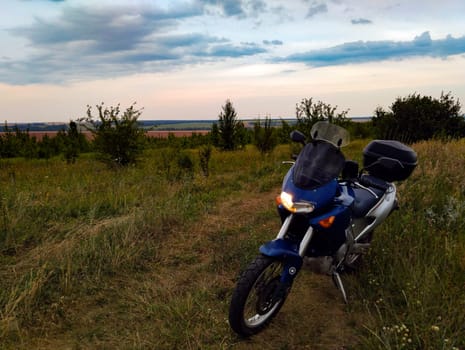 A motorcycle rests on grassy terrain as the sun sets, casting warm hues over the landscape with fields in the distance.
