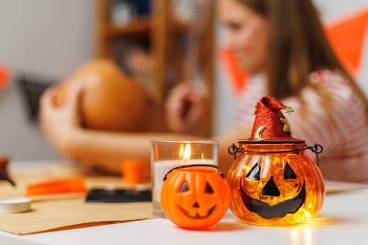 Woman in striped shirt creating Halloween pumpkin lantern on decorated table with candle and accessories. Studio craft session.