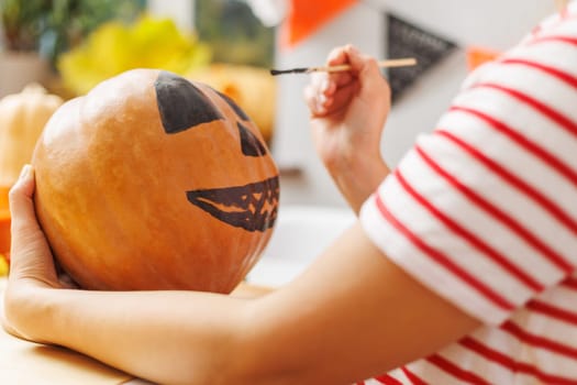 Close-up of hands painting a scary face on an orange pumpkin. Close-up photograph with selective focus. Halloween preparation concept. Design for invitation, poster.