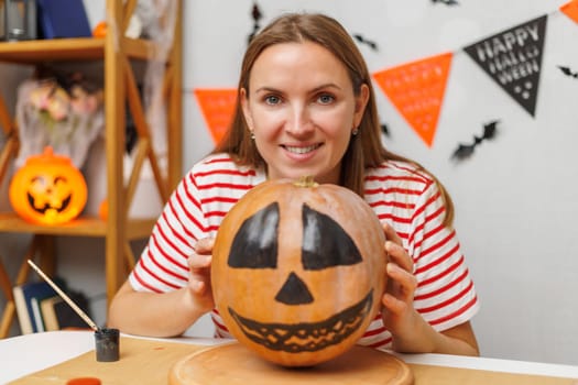 Adult female holding a painting pumpkin with smiley face, wearing striped shirt. Halloween preparation and decoration concept. Suitable for banner, invitation, poster.