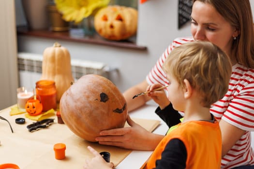 Mother and young child together painting a pumpkin at a decorated Halloween craft table. Family bonding and Halloween preparation concept for design and greeting.