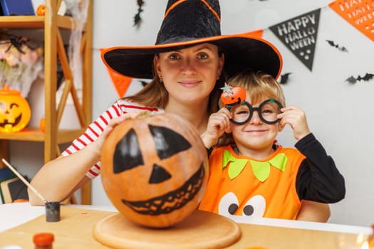 Mother and child in Halloween costumes decorating a pumpkin. Studio portrait with decorations. Halloween family fun concept. Design for greeting card, banner.