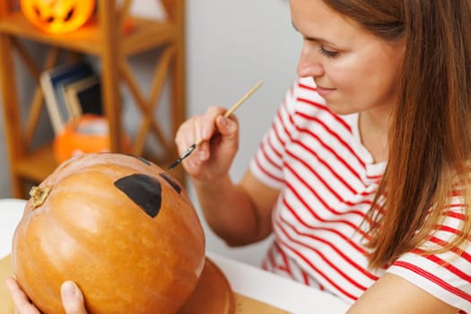 Adult woman painting a black pattern on an orange pumpkin. Indoor casual candid scene. Halloween decoration and crafting concept for design and poster.