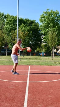 An elderly old Italian man plays basketball on a sports court on a summer day. Vertical