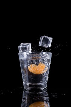 A studio photo of ice cubes being dropped into a glass of water with an orange slice.