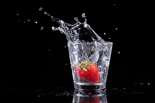 studio photo of strawberries and ice dropped into a water glass against a black background.