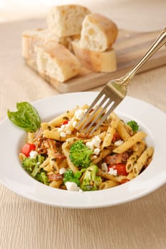 Studio photo of a fork going into a bowl of penne pasta with broccoli and feta cheese and a cutting board of ciabatta bread slices.