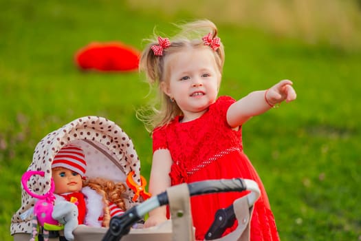 A cheerful girl wearing a bright red dress is happily pointing while she pushes a stroller with her doll outdoors