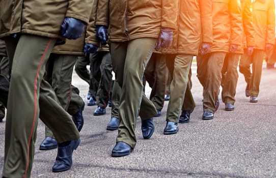 A group of soldiers dressed in green military uniforms march together in formation on a parade ground during a military ceremony. They are wearing black gloves and their faces are not visible.