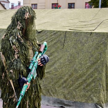 A soldier dressed in camouflage holds a rifle, standing near a green tent.