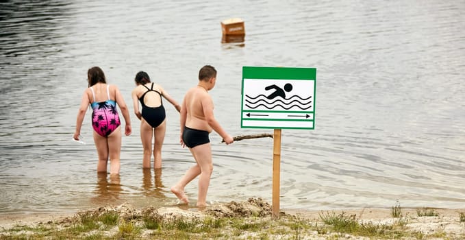 Three children play by the water's edge, laughter filling the air as they wade into the lake on a bright, sunny day. Two girls, in colorful swimsuits, explore shallow waters, while a boy, holding a stick, gazes towards a clearly marked swimming safety sign.