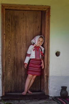 A young girl dressed in a stunning and elegant traditional dress poses gently by a rustic wooden door