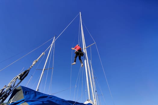 Man working up the mast of a sailing yacht, climbing with rope and bosun's chair on a sunny day with blue sky, repairing, fixing the mast shrouds