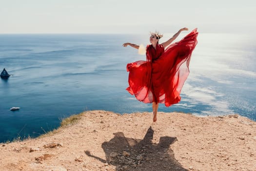 Side view a Young beautiful sensual woman in a red long dress posing on a rock high above the sea during sunrise. Girl on the nature on blue sky background. Fashion photo.