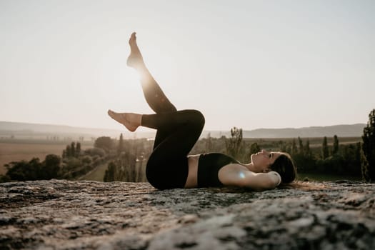 Well looking middle aged woman with long hair, fitness instructor in leggings and tops doing stretching and pilates on the rock near forest. Female fitness yoga routine concept. Healthy lifestyle.