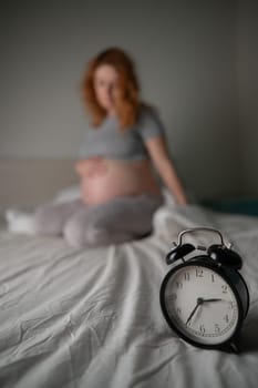 Caucasian pregnant woman sits on the bed and suffers from insomnia. Alarm clock in the foreground