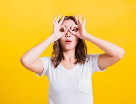 Portrait Asian Thai beautiful young woman standing, She made finger OK symbol sign to agree near two eyes and looking at camera, shoot photo in studio on yellow background, There was copy space