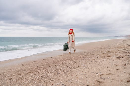 Redhead woman Christmas tree sea. Christmas portrait of a happy redhead woman walking along the beach and holding a Christmas tree in her hands. She is dressed in a light coat and a red beret