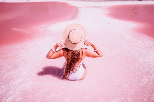 A woman traveler looks at an amazing pink salt lake. He sits with his back in a bathing suit and holds his hat in his hands. Wanderlust travel concept.