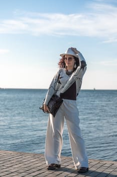 Stylish seashore woman. Fashionable woman in a white hat, white trousers and a light sweater with a black pattern on the background of the sea