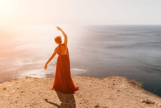 Side view a Young beautiful sensual woman in a red long dress posing on a rock high above the sea during sunrise. Girl on the nature on blue sky background. Fashion photo.