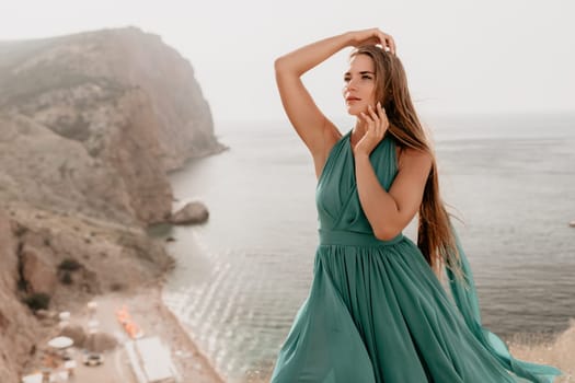 Side view a Young beautiful sensual woman in a mint long dress posing on a volcanic rock high above the sea during sunset. Girl on the nature on overcast sky background. Fashion photo