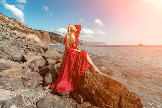 Red dress sea woman. A blonde with flowing hair in a long red dress sits on a rock near the sea. The concept of trips, a photo shoot at the sea.